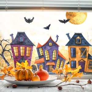 Op de afbeelding: Een raam met een Halloween-thema met drie cartoonhuizen, vleermuizen en een volle maan. De huizen zijn versierd met oranje, paarse en gele verf. Er liggen pompoenen en herfstbladeren op de vensterbank.
