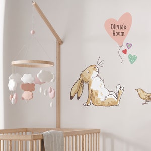 May include: A nursery wall decal set featuring a brown bunny rabbit with a pink heart balloon that says "Olivia's Room" and a small brown bird. The decal set is on a white wall with a pink and white cloud mobile hanging from the ceiling.