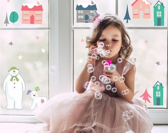 May include: A young person in a pink dress blowing bubbles near a window decorated with winter-themed stickers. The stickers include houses, trees, snowflakes, stars, a snowman, and a dog.