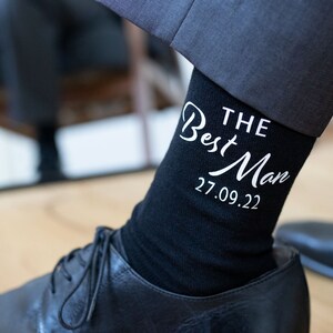 Peut inclure: Chaussettes noires avec un texte blanc qui dit "THE Best Man 27.09.22".