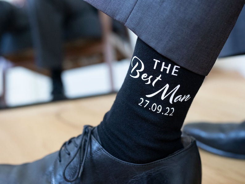 Peut inclure: Chaussettes noires avec un texte blanc qui dit "THE Best Man 27.09.22".