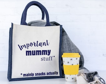 May include: A white canvas tote bag with navy blue trim and handles. The bag has the text "Important Mummy Stuff* *mainly snacks actually" printed on it. A yellow reusable coffee cup with a bee pattern is sitting in front of the bag.