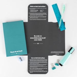 May include: A set of iron-on and stick-on name labels for clothing and other items. The labels are packaged in a black folder with instructions on how to use them. The folder also includes a ruler, a pencil sharpener, paper clips, and a highlighter.