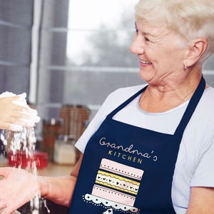 Op de afbeelding: Een vrouw draagt een blauwe schort met de tekst "Grandma's Kitchen" en een roze en geel taartontwerp. Ze glimlacht en kijkt naar rechts. Een hand houdt een handvol wit poeder vast, dat naar beneden valt.