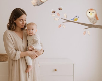 May include: A woman holding a baby in a beige dress, standing in front of a white dresser. The wall features whimsical illustrations of owls, bees, and a blue bird perched on a branch with leaves, creating a nursery theme.