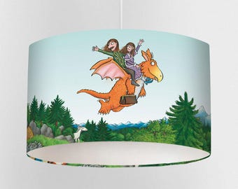 May include: A cylindrical lampshade featuring a whimsical illustration of two children riding an orange dragon through a landscape of green trees and mountains under a light blue sky. The dragon has pink wings and a brown suitcase.