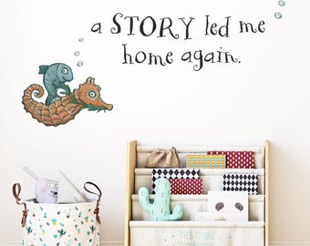 May include: A whimsical wall decal featuring a seahorse and fish illustration, with the text "a STORY led me home again." A wooden bookshelf and a cactus-themed storage bin are also visible.
