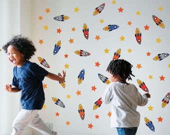 May include: Two children play in a room decorated with colorful rocket and star wall decals. The decals are blue, red, yellow, and orange. The children are wearing casual clothing and smiling.