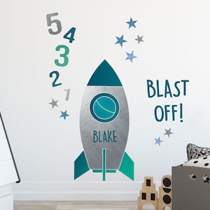 May include: A silver and teal rocket ship wall decal with the name "Blake" on it. The decal also includes the words "Blast Off!" and a set of numbers 1 through 5. There are also several silver and blue glitter stars.