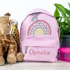 May include: Pink backpack with a rainbow design and the name "Ophelia" embroidered on the front. A stuffed giraffe toy, a water bottle, and a pink lunchbox are also in the image.