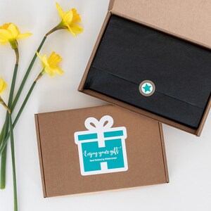 May include: A brown cardboard box with a teal gift box sticker that says "Enjoy your gift" and a black star sticker. The box is open and a black gift bag is inside.