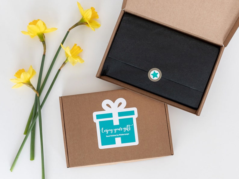 May include: A brown cardboard box with a teal gift box sticker that says "Enjoy your gift" and a black star sticker. The box is open and a black gift bag is inside.