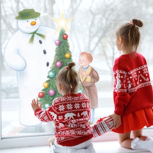 May include: Two young children in red sweaters with white snowflake and bear patterns look at a Christmas scene on a window. The scene includes a snowman, a decorated Christmas tree, and a boy in a coat. One child holds a wrapped gift.