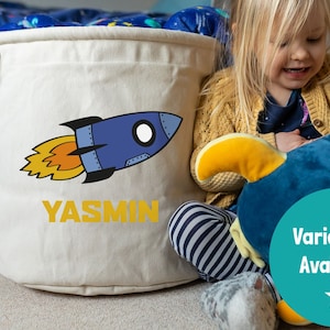 May include: A white canvas storage bin with a blue rocket ship and orange flames design. The bin has a drawstring closure and the name "YASMIN" is printed in yellow letters. A child is sitting next to the bin holding a blue rocket ship plush toy.