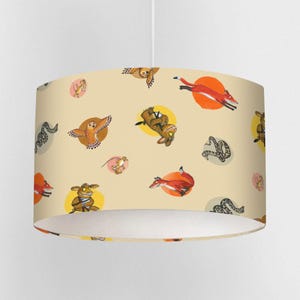 Könnte beinhalten: Zylindrischer Lampenschirm mit einem sich wiederholenden Muster von Cartoon-Waldtieren, darunter Füchse, Eulen und Schlangen, auf hellbeigem Hintergrund. Die Illustrationen sind in gelben und rosa Kreisen angeordnet.