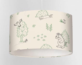 May include: Cream-colored lampshade featuring a whimsical forest scene with green line drawings. The design includes trees, birds, a rabbit, and a creature holding a book. The lampshade is cylindrical and suspended from a white cord.