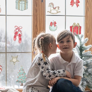 Puede incluir: Dos niños, una niña y un niño, están sentados junto a una ventana decorada con pegatinas de temática navideña. La niña le susurra al oído al niño. Un pequeño árbol de Navidad artificial está en primer plano.