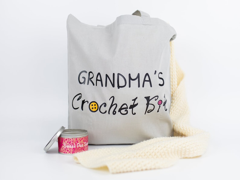 May include: A gray canvas tote bag with the text "Grandma's Crochet Bit" in black lettering and a yellow button graphic. A white crocheted scarf and a pink and silver tin with the text "Sweet Pea Soap" are also in the image.