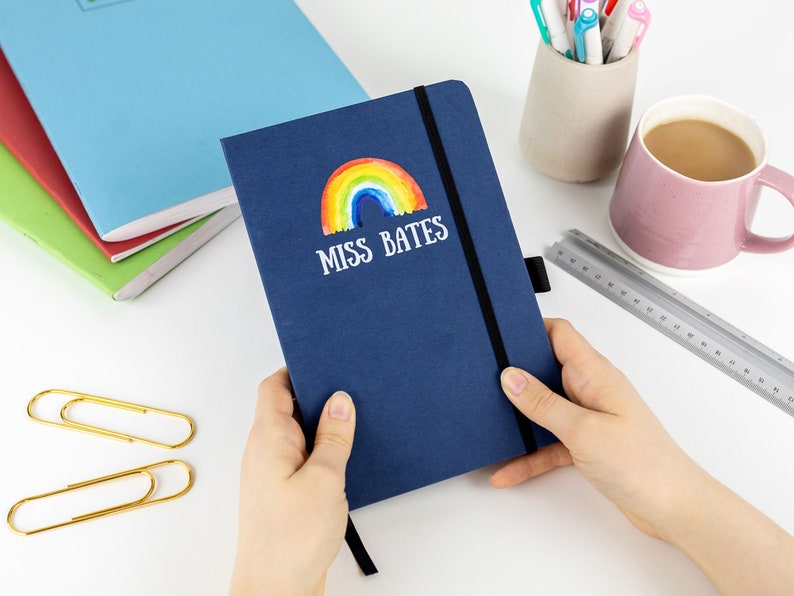 Puede incluir: Una libreta azul marino con un arco&iacute;ris y el texto "MISS BATES" en la portada. La libreta est&aacute; siendo sostenida por las manos de una persona.