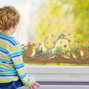 May include: A child looks out a window at a colorful window cling featuring a worm, frogs, and other insects. The cling is on a window with a white frame.
