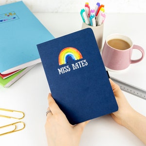 Puede incluir: Una libreta azul marino con un arco&iacute;ris y el texto "Miss Bates" en la portada.