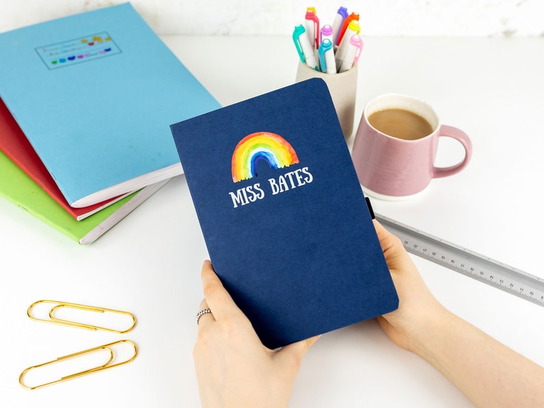 Puede incluir: Una libreta azul marino con un arco&iacute;ris y el texto "Miss Bates" en la portada.