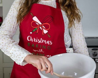Puede incluir: Delantal rojo para hornear de Navidad con un rodillo y un batidor blancos atados con un lazo rojo. El texto en el delantal dice "Christmas Baking with Elaine".