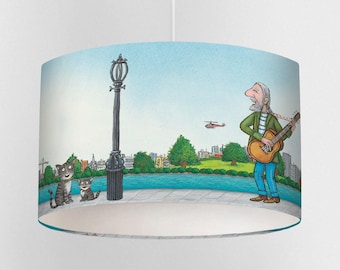 May include: A cylindrical lampshade featuring a whimsical illustration. The design depicts a man playing a guitar near a body of water, with two cats and a lamppost. A helicopter flies in the sky. The background shows a cityscape under a blue sky.