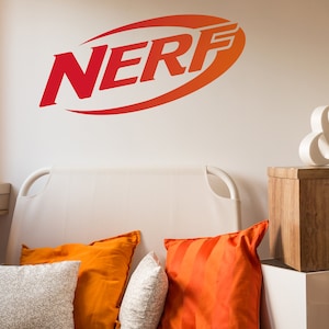 Puede incluir: Un sofá blanco con almohadas a rayas naranja y blancas y una almohada blanca con un patrón gris. El sofá está frente a una pared blanca con un logotipo "NERF" rojo y naranja. Un signo de ampersand de madera blanca se encuentra en una caja de madera.