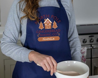 Puede incluir: Un delantal azul con un diseño de casa de jengibre y árboles. El texto dice "North Pole Gingerbread Baking Co. Natalie".