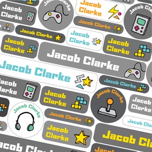 Könnte beinhalten: Ein Satz bunter, personalisierter Namensschilder mit dem Namen "Jacob Clarke" in verschiedenen Schriftarten und Stilen. Die Etiketten enthalten Bilder von Videospielcontrollern, Sternen und Pixelblöcken.