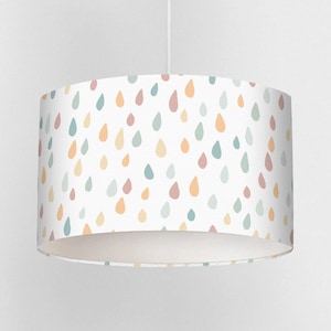 Puede incluir: Pantalla de lámpara cilíndrica con fondo blanco y un patrón de gotas de agua de colores pastel en tonos azules, naranjas, amarillos y rosas. La pantalla cuelga de un cable blanco.