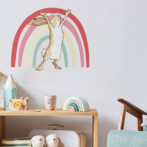 May include: A beige bunny rabbit with long ears stands on its hind legs with its arms raised above its head. The bunny is in front of a multi-colored rainbow with pink, red, yellow, and green stripes.