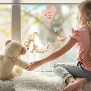 May include: A young girl sits by a window with a teddy bear. The window is decorated with a heart-shaped balloon that says "Lucie's Birthday Party" and other heart-shaped decorations.