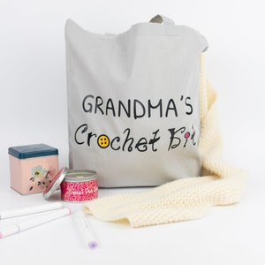 May include: A gray canvas tote bag with the text "Grandma's Crochet Kit" in black lettering. The bag is filled with a variety of crochet supplies, including yarn, needles, and a small tin.