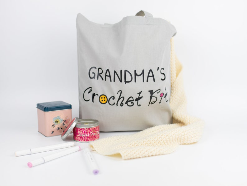 May include: A gray canvas tote bag with the text "Grandma's Crochet Kit" in black lettering. The bag is filled with a variety of crochet supplies, including yarn, needles, and a small tin.