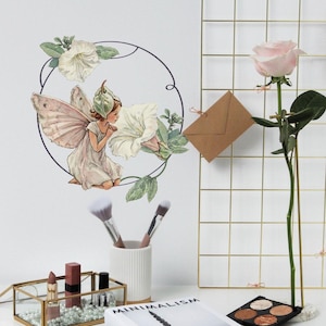 Puede incluir: Un hada blanca con alas rosas y un sombrero verde se representa en un marco circular con flores blancas y hojas verdes. El hada está mirando una flor blanca. La imagen está sobre un fondo blanco.