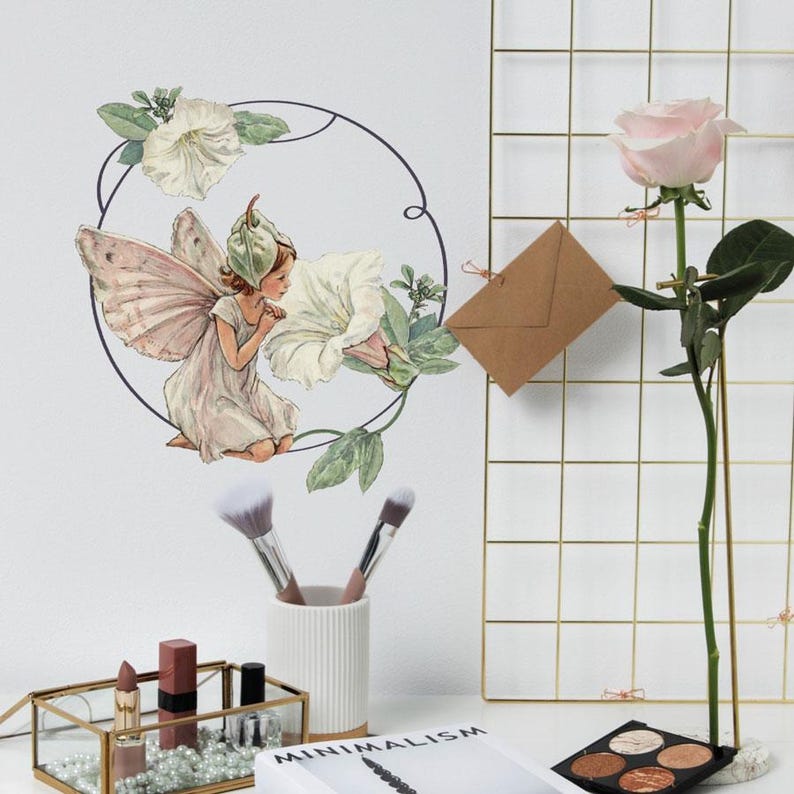 May include: A white fairy with pink wings and a green hat is depicted in a circular frame with white flowers and green leaves. The fairy is looking at a white flower. The image is on a white background.