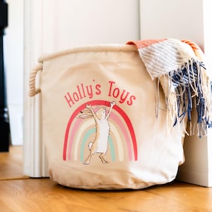 May include: Beige canvas toy storage bin with rope handles. The bin features a rainbow and a cartoon rabbit graphic with the text "Holly's Toys". A colorful blanket with fringe is draped over the top of the bin.