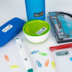May include: A set of colourful personalised labels with the name "Adam Williams" printed on them. The labels are designed with a variety of sea creatures, including a dolphin, a manta ray, and a shark. The labels are shown on a variety of items, including a water bottle, a lunchbox, a pencil case, a marker, and a pair of glasses.