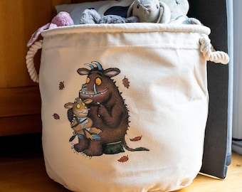 May include: A cream-colored canvas storage bin with a drawstring closure and rope handles. The bin features a colorful illustration of two cartoon monsters and falling leaves. The bin is filled with stuffed animals and is sitting on a wooden floor.
