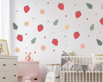 May include: A white wall decorated with red strawberries, pink flowers, green leaves, and red and green dots. A white crib with a gray and pink banner is in the foreground. A pink storage box and a white dresser are also visible.
