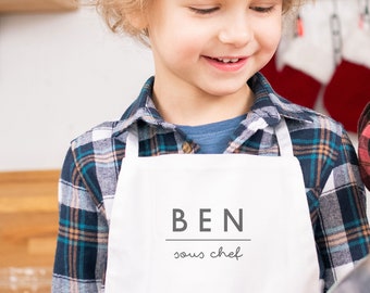 Könnte beinhalten: Eine weiße Schürze mit dem Namen "BEN" und dem Text "sous chef" darauf gedruckt. Ein kleines Kind trägt die Schürze.