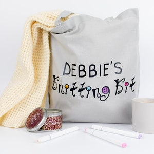May include: A gray canvas tote bag with the text "DEBBIE'S Knitting Kit" printed on it. A yellow knitted scarf, a small tin candle, and three white markers are also in the image.