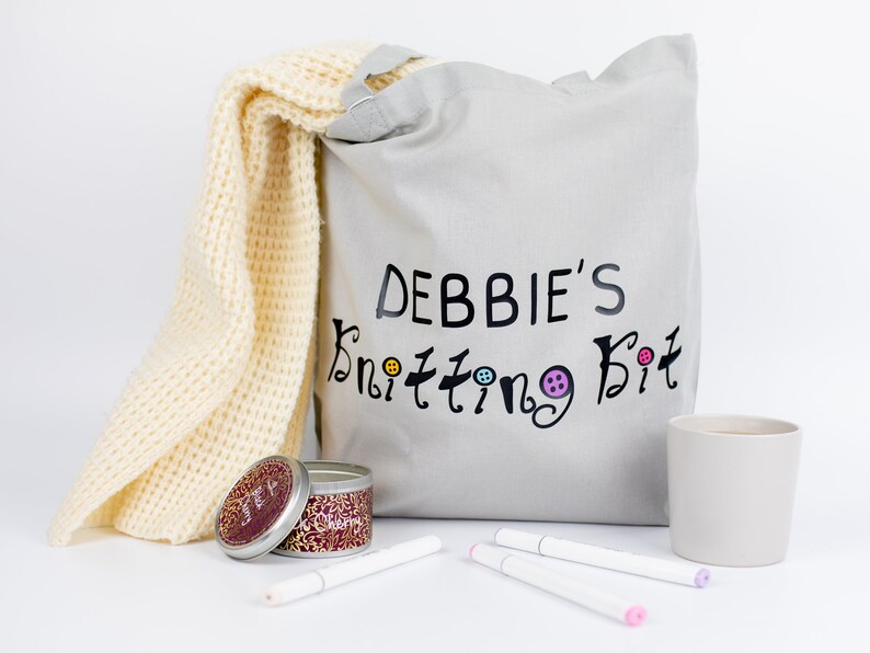 May include: A gray canvas tote bag with the text "DEBBIE'S Knitting Kit" printed on it. A yellow knitted scarf, a small tin candle, and three white markers are also in the image.