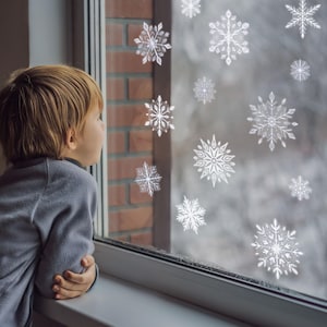 以下が含まれることがあります： 白い雪の結晶のステッカーで飾られた窓から、子供が見ています。窓は白い窓枠とレンガの壁で囲まれています。背景には、雪景色がぼやけています。