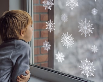May include: A young person gazes out a window adorned with decorative white snowflake decals. The window is framed by a white sill and a brick wall. The background shows a snowy, blurred outdoor scene.