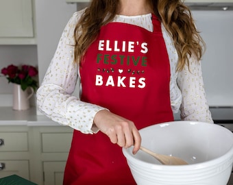 Puede incluir: Un delantal rojo con el texto "Ellie's Festive Bakes" en blanco con acentos verdes. El delantal lo lleva puesto una persona en un entorno de cocina.