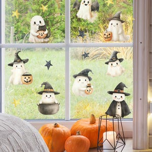 May include: A window decorated with Halloween-themed stickers featuring cartoon ghosts wearing witch hats and holding jack-o'-lanterns. Orange pumpkins and a candle holder are in front of the window, creating a festive autumn scene.