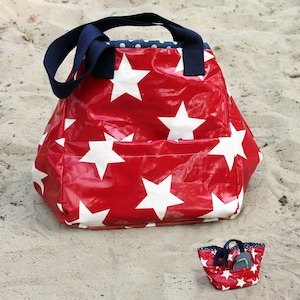 May include: A red tote bag with white star patterns and navy blue handles sits on sand. A smaller matching bag is next to it. The bags have a pocket on the front and a blue and white polka dot lining.
